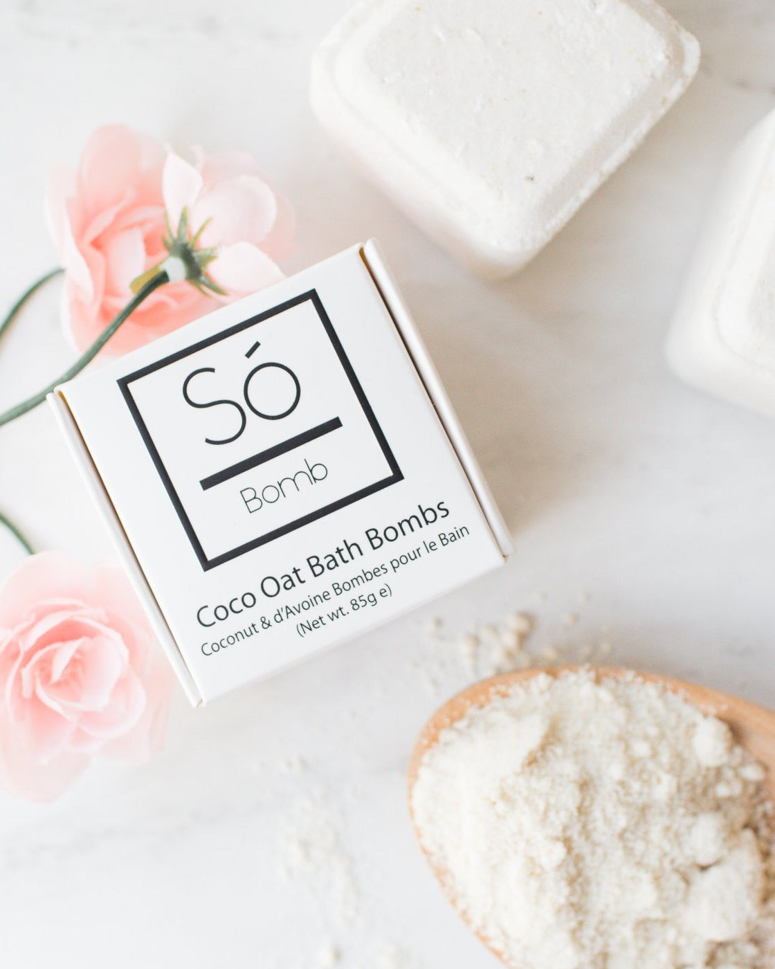 A styled flat-lay of a white box of Só Bomb Coco Oat Bath Bombs on a light marble surface, surrounded by pink roses, finished bath bombs, and a wooden spoon filled with the white powder.