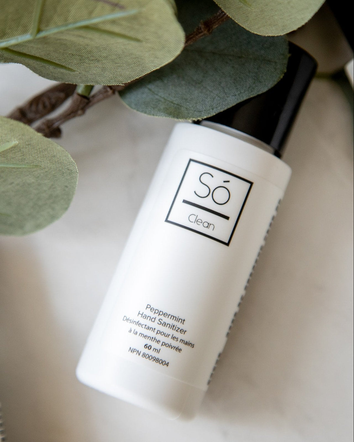 A white bottle of Só Clean peppermint hair sanitizer with a black cap, styled on a light surface with green leaves in the foreground.