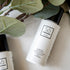 A white bottle of Só Clean peppermint hair sanitizer with a black cap, styled on a light surface with green leaves in the foreground.