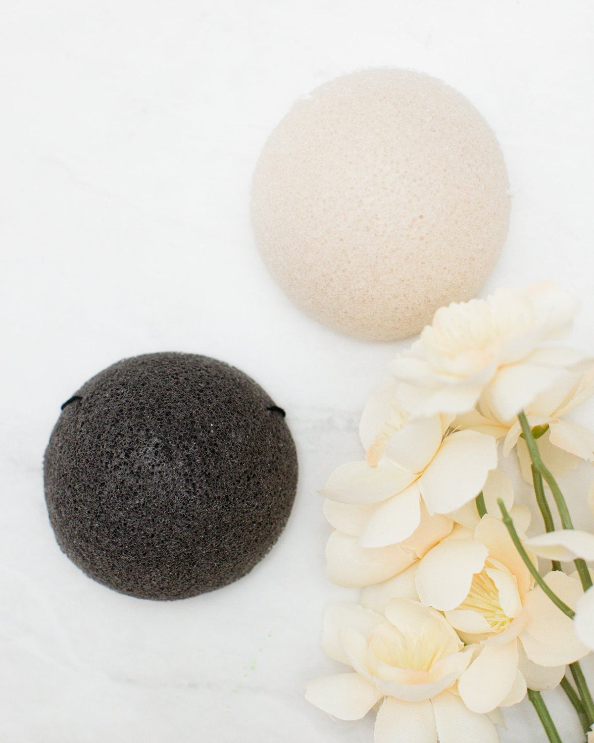 Two round konjac sponge, one cream coloured and one dark charcoal gray, arranged on a white marble surface next to cream-colored flowers.