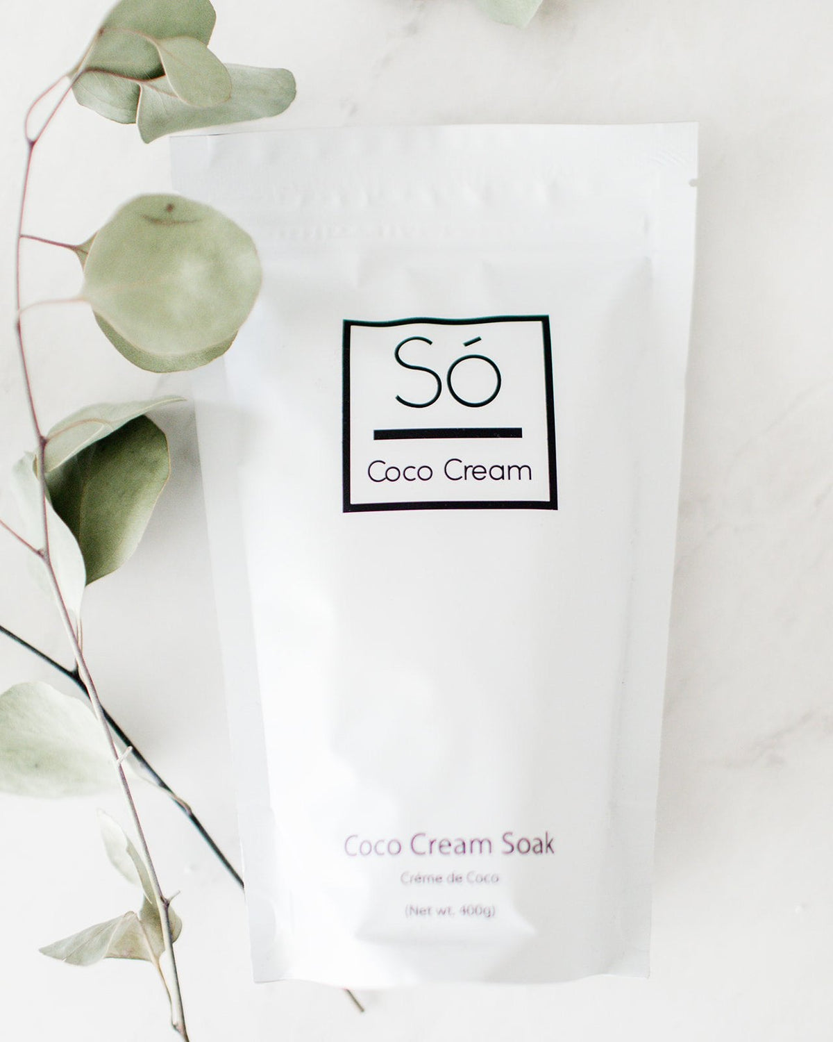 A packaged coconut milk bath soak, labeled 'Só Luxury Coco Cream Soak' on a marble surface with a leafy green plant stem.