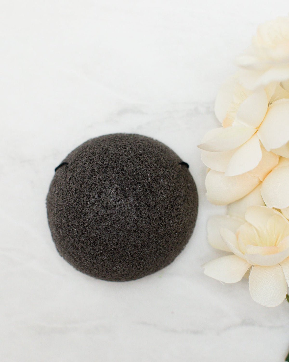 A dark charcoal gray coloured round konjac sponge arranged on a white marble surface next to cream-colored flowers.