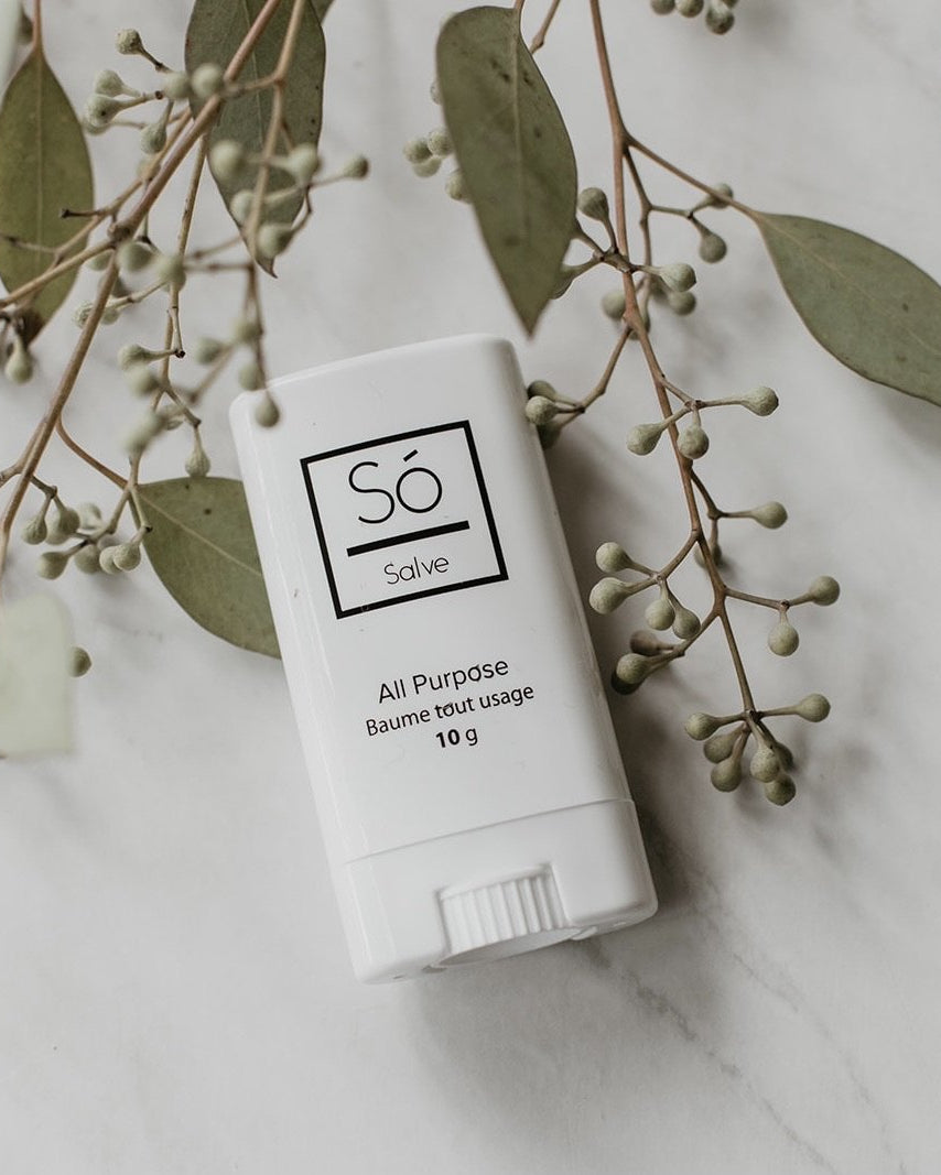 A white plastic deodorant style container branded Só Salve All purpose healing salve in 10 g twist-up stick styled on a marble background with greenery.