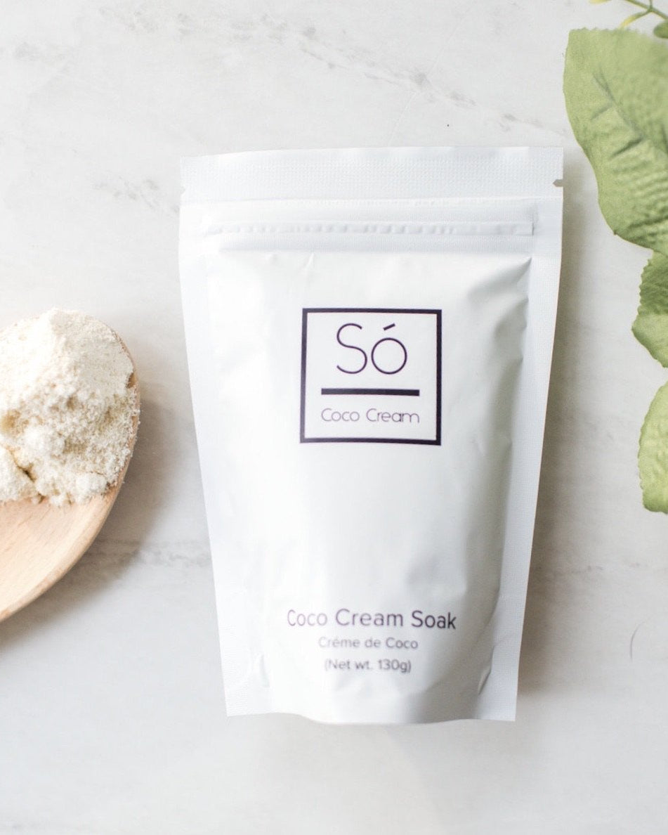 A white stand-up pouch labeled 'Só Coco Cream' and 'Coco Cream Soak' on a light marble surface, styled with a wooden spoon of white powder and green leaves.
