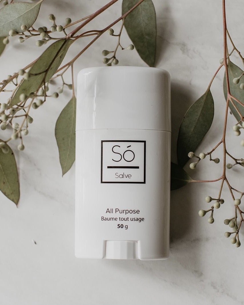 A white plastic deodorant style container branded Só Salve All purpose healing salve in 50 g twist-up stick styled on a marble background with greenery.